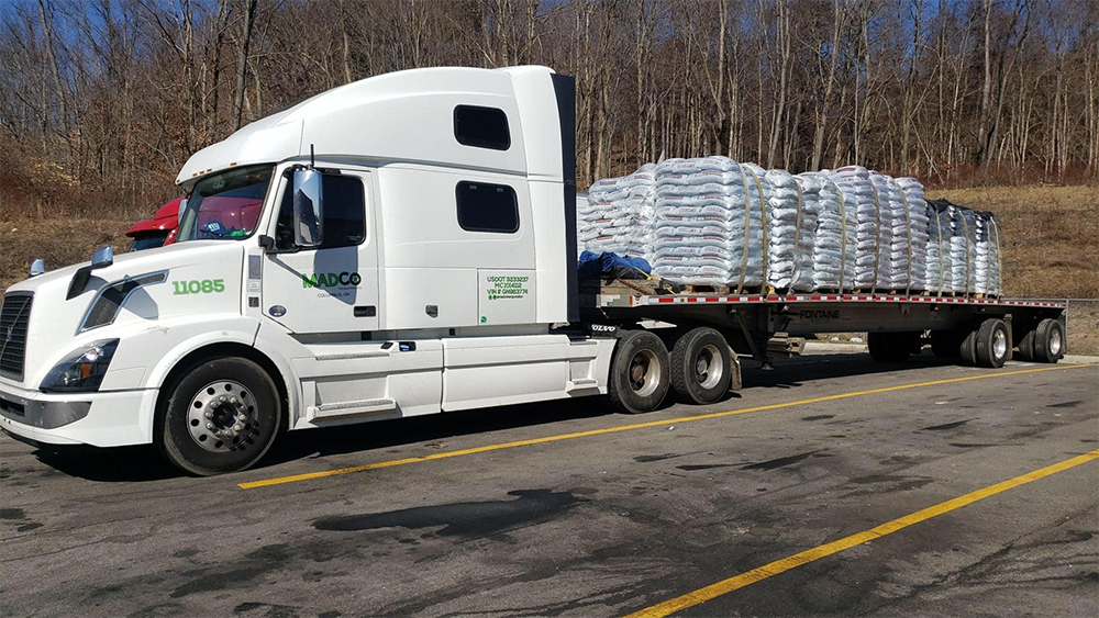 A large white semi-truck is parked on a highway, carrying a flatbed trailer loaded with bags stacked neatly.