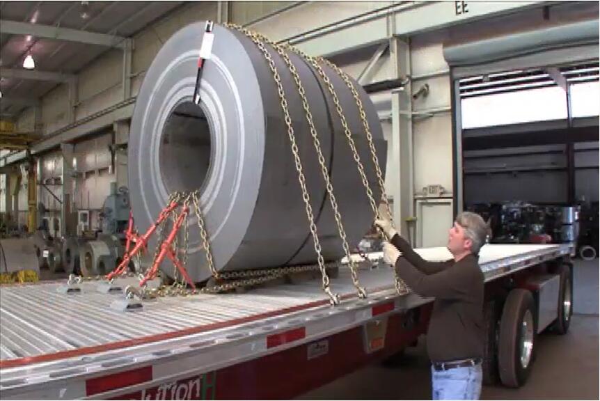 A large metal coil is being secured onto a truck trailer using heavy chains and a lifting mechanism.
