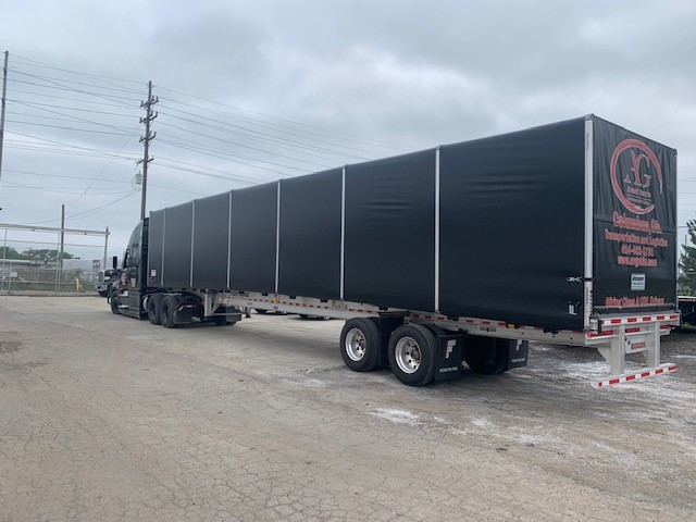 A large black semi-truck with a covered trailer is parked on a gravel lot under an overcast sky.