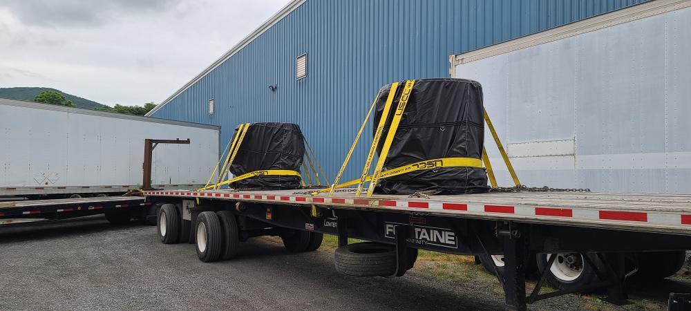 Two large objects are secured on a flatbed trailer, covered in black tarps and strapped down with yellow belts, parked next to a blue building.