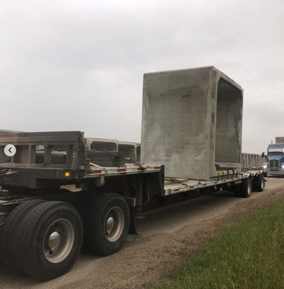 A flatbed truck carries a large, rectangular concrete structure along a road under an overcast sky.