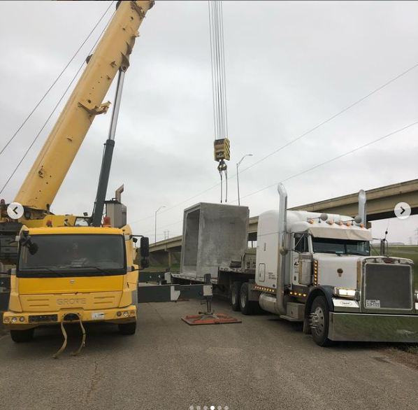 A yellow crane is lifting a large concrete structure from a flatbed truck on a construction site beneath an overpass.