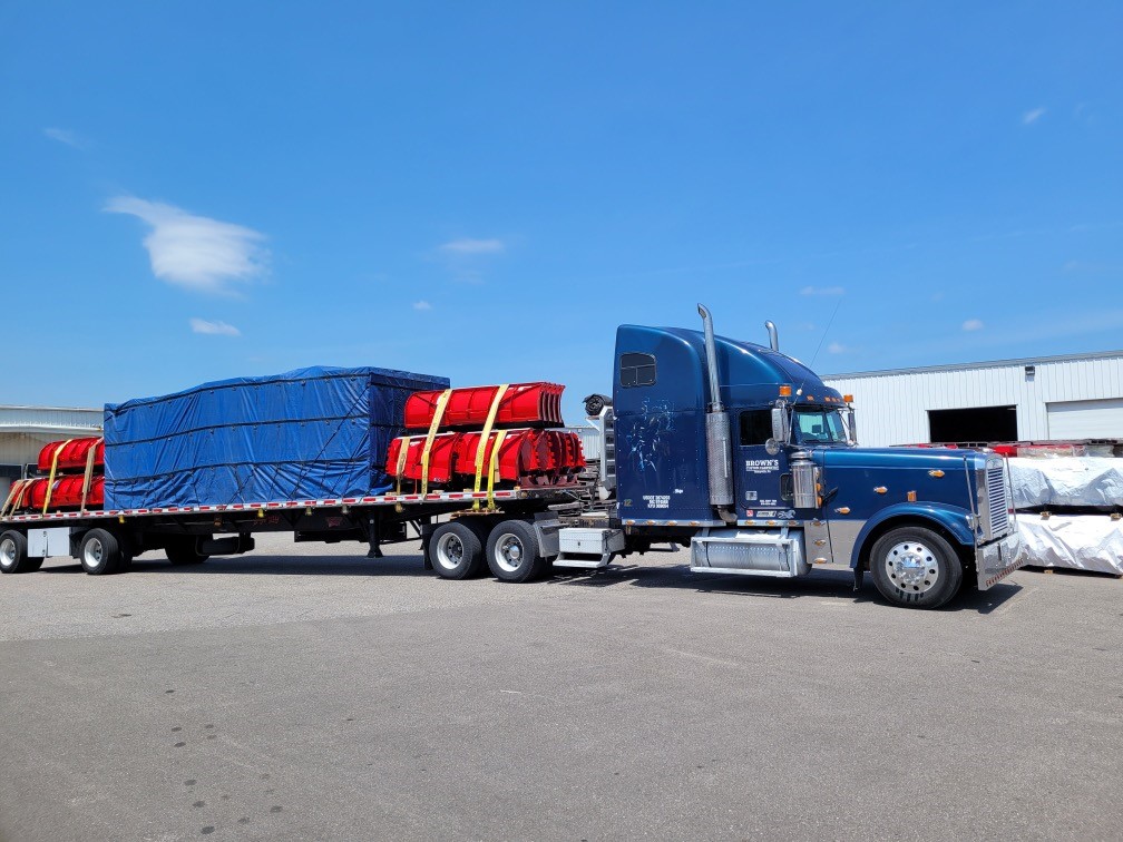 A blue truck is parked with a large trailer covered in a blue tarp, carrying several red items secured with straps against a clear sky.