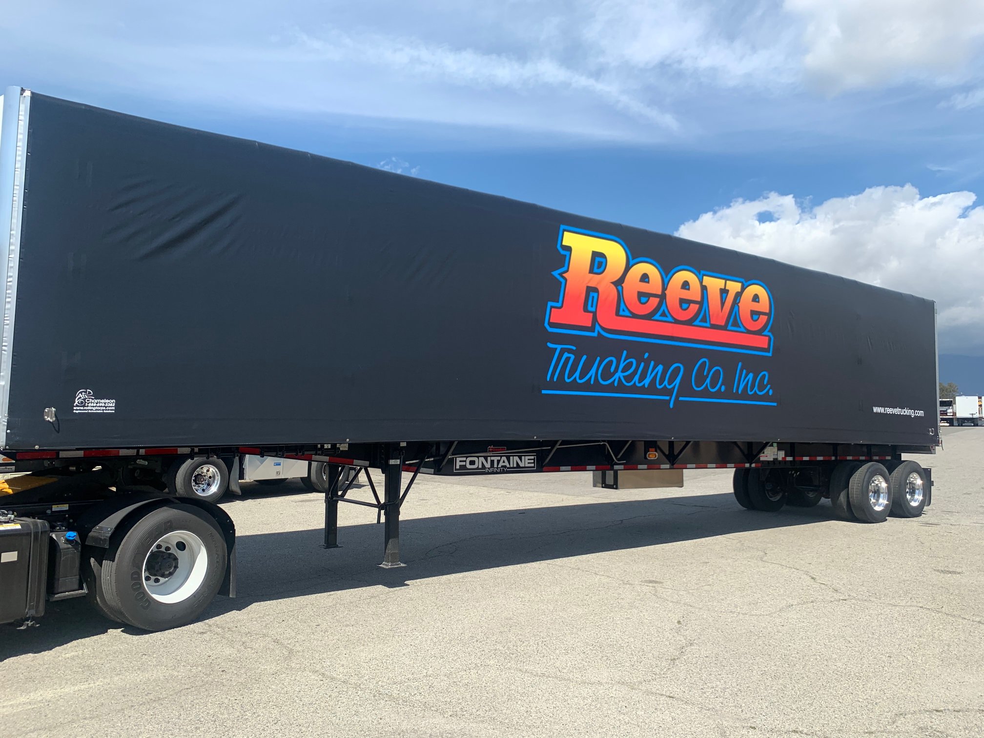 A large black trailer with a colorful logo for Reeve Trucking Co. Inc. is parked on a paved surface under a partly cloudy sky.