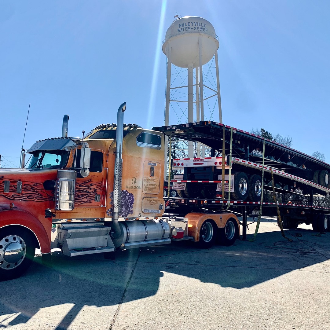 A brightly colored truck with flame designs pulls a long flatbed trailer beneath a large water tower.