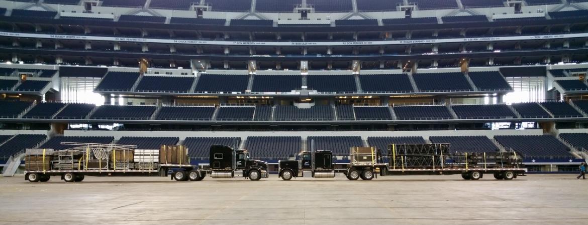 Large trucks loaded with equipment are lined up on a vast, empty sports arena floor with tiered seating in the background.