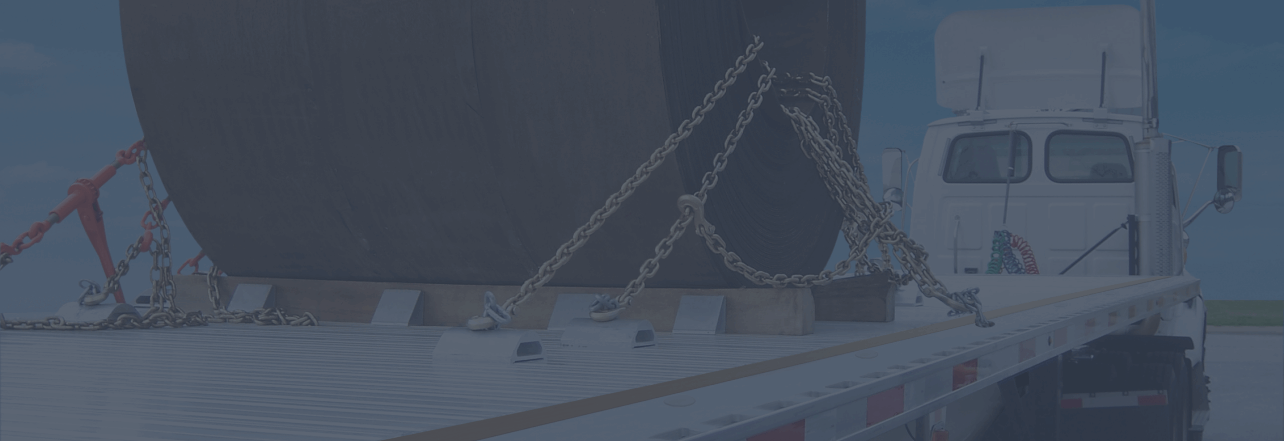 A large cylindrical object is secured on a flatbed truck with chains in a bright outdoor setting.