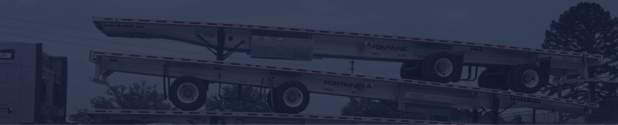 A heavy-duty trailer with multiple levels is parked, showcasing its metallic structure and wheels against a backdrop of trees and power lines.